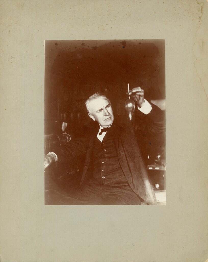 Thomas Edison holding a small electric lamp or test tube in his laboratory in 1905